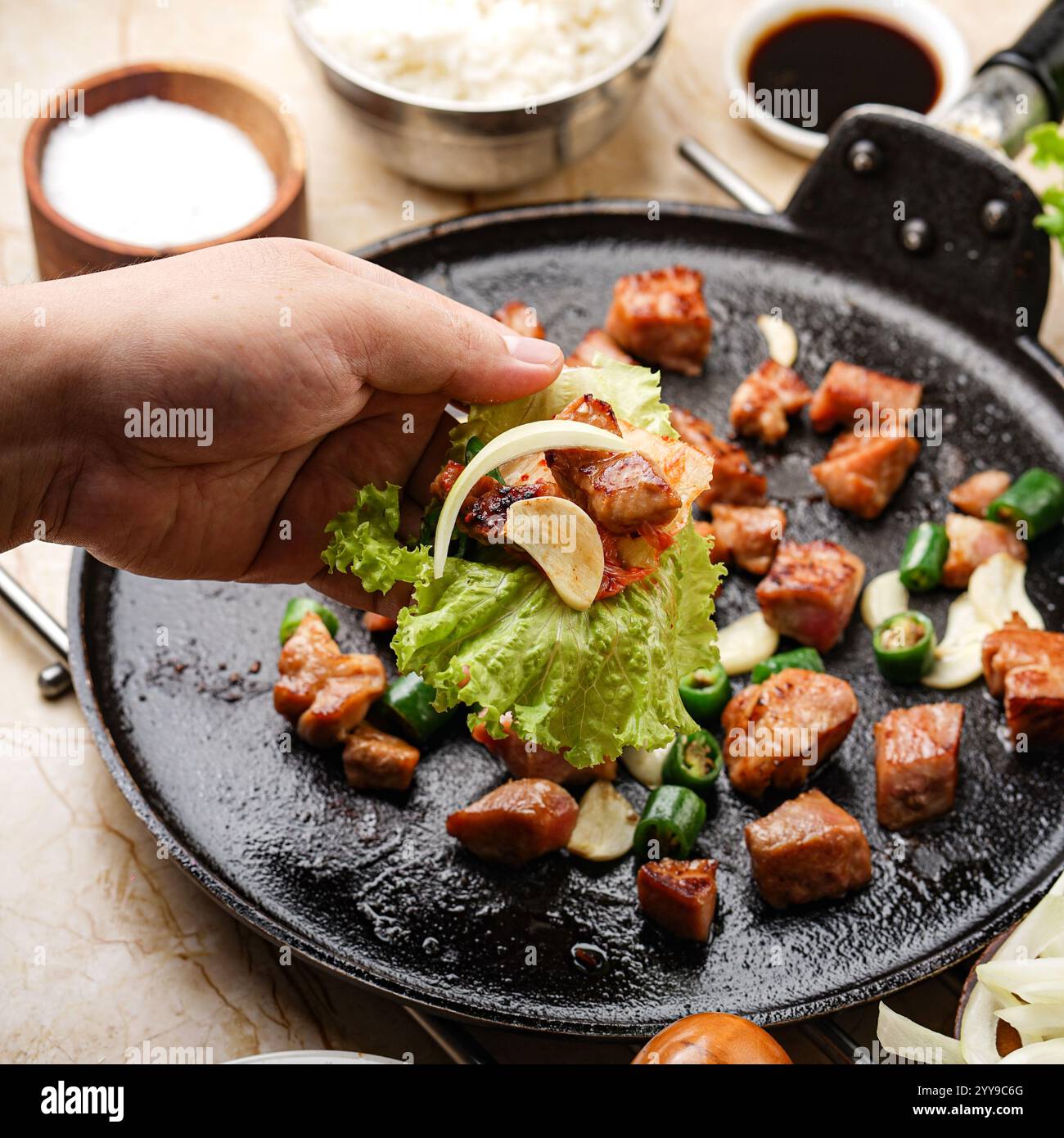 Pancetta di maiale grigliata servita sfrigolante su una padella di ghisa, avvolta in lattuga fresca con aglio, cipolla e peperoncino per un perfetto stile coreano Foto Stock