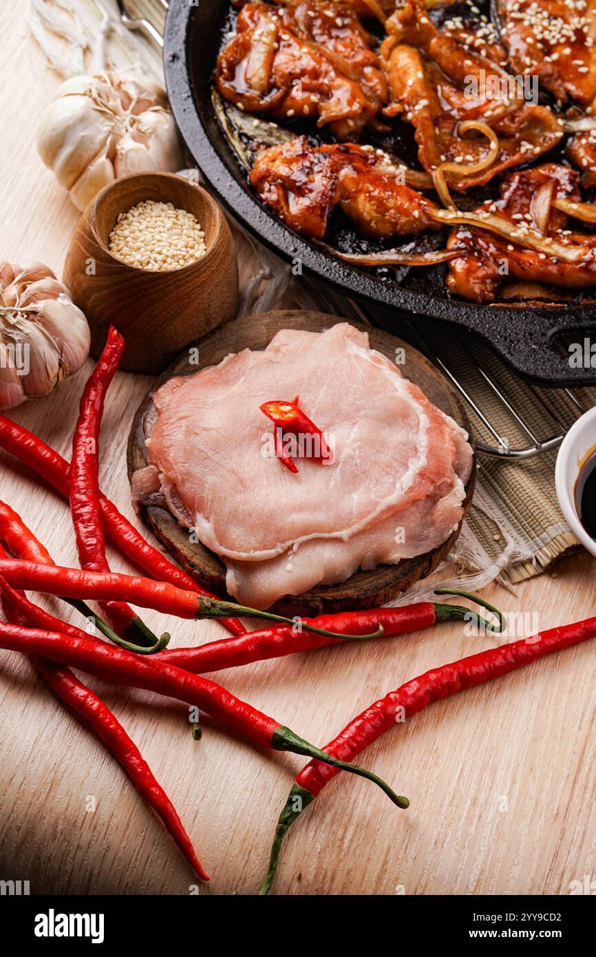 Fette di maiale fresche circondate da peperoncini rossi, aglio e semi di sesamo, ingredienti pronti per essere trasformati in una salata frittura speziata, con un Foto Stock