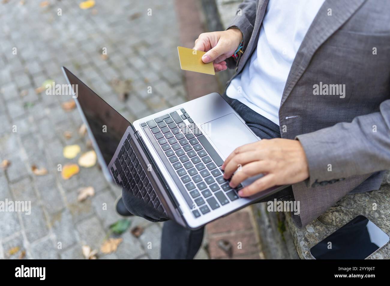 Vista ravvicinata dall'alto di un uomo d'affari irriconoscibile che usa la carta di credito per fare acquisti online all'aperto seduto su una panchina Foto Stock