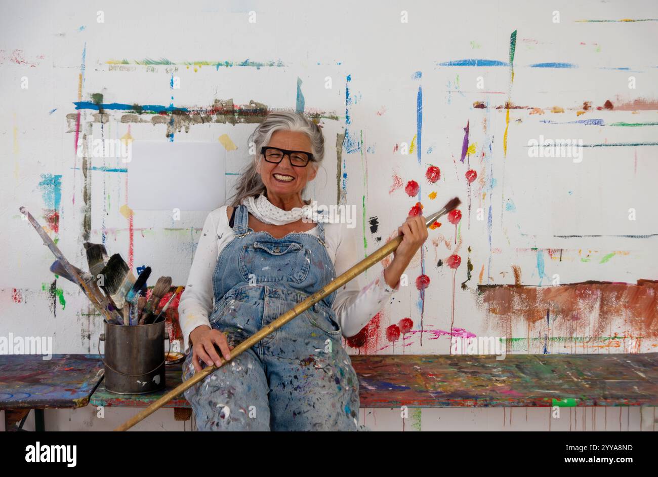 Maturo artista donna maturo, pazzo, capelli e occhiali grigi si siede gioiosamente su una parete colorata, tenendo una grande spazzola. Le sue tute in denim riflettono un Foto Stock
