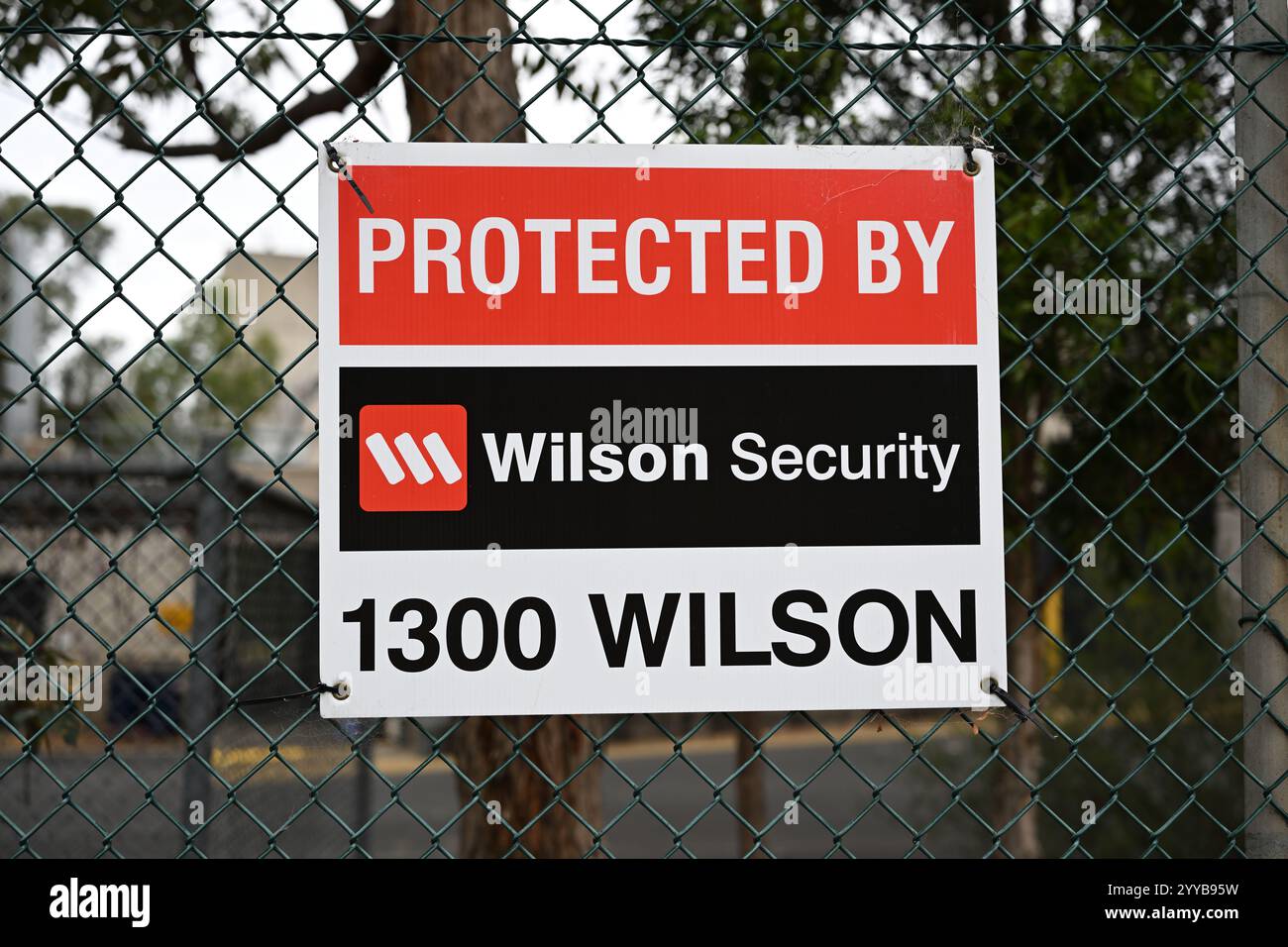 Protetto dal cartello Wilson Security con il logo dell'azienda, su una recinzione a catena, con edifici e alberi sullo sfondo Foto Stock