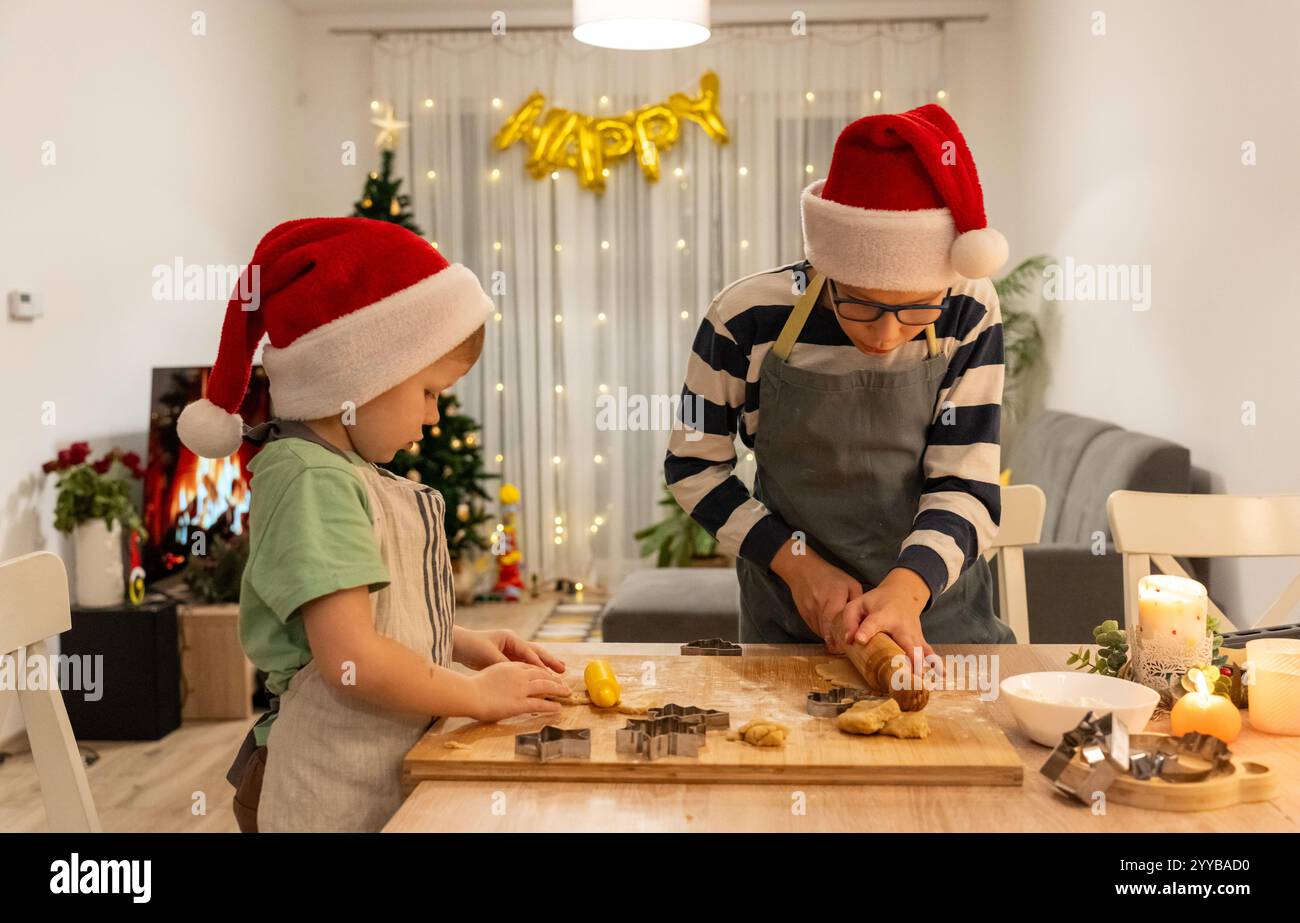 Due bambini felici che indossano cappelli di Babbo Natale preparano i biscotti di Natale in una calda cucina festosa piena di decorazioni natalizie e luci accese. Famiglia Foto Stock