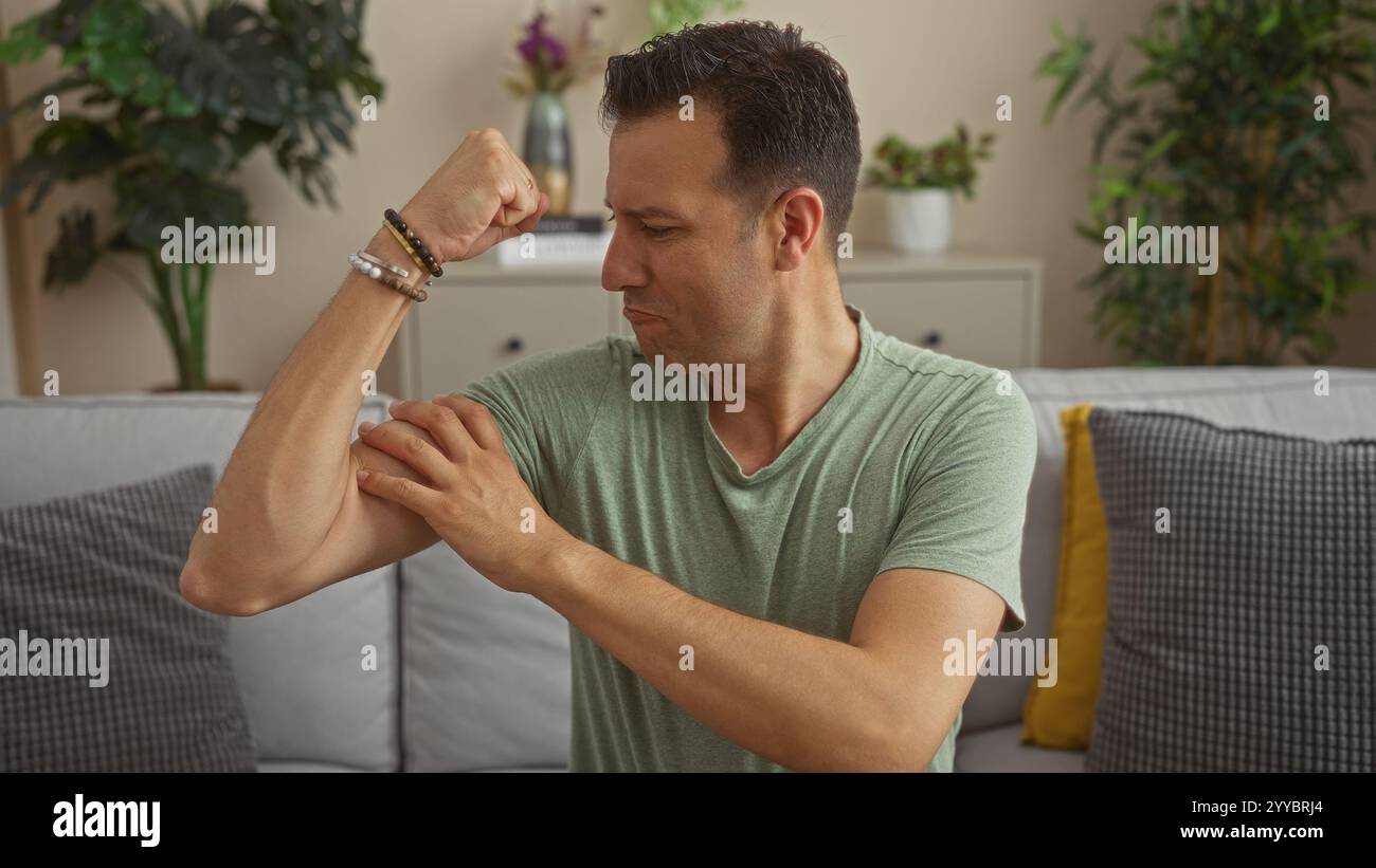 Uomo ispanico con una camicia verde che si flette i muscoli mentre si siede in un accogliente soggiorno con piante e cuscini intorno, mostrando forza in casa. Foto Stock