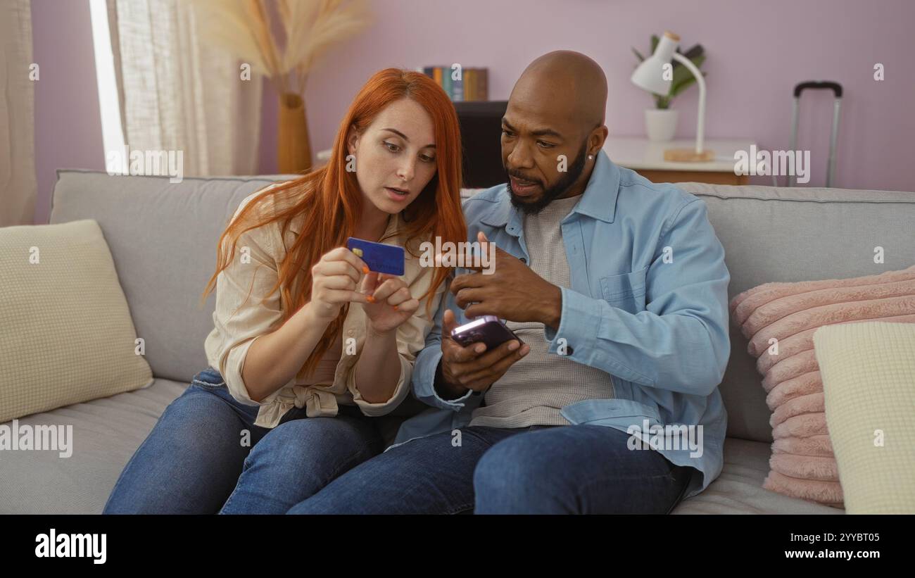Coppia interrazziale seduta insieme in un soggiorno con una carta di credito e uno smartphone, discutendo di shopping online o di banca, evidenziando un moder Foto Stock
