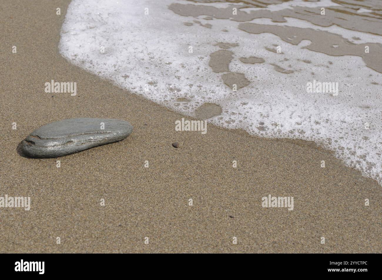 Una spiaggia tranquilla con una superficie sabbiosa di colore chiaro e dolci onde che colpiscono la riva. Una sola pietra si trova nella sabbia, in Spagna, in Europa Foto Stock