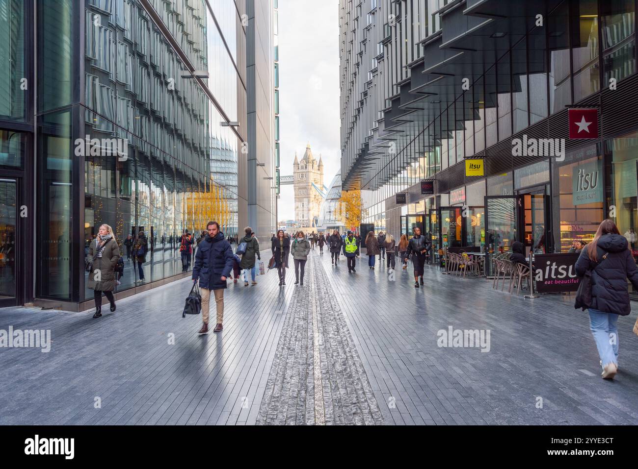 Londra, Regno Unito - 3 dicembre 2024: Persone che camminano lungo un passaggio attraverso i moderni edifici di uffici a Londond Bridge City. Tower Bridge è sullo sfondo. Foto Stock