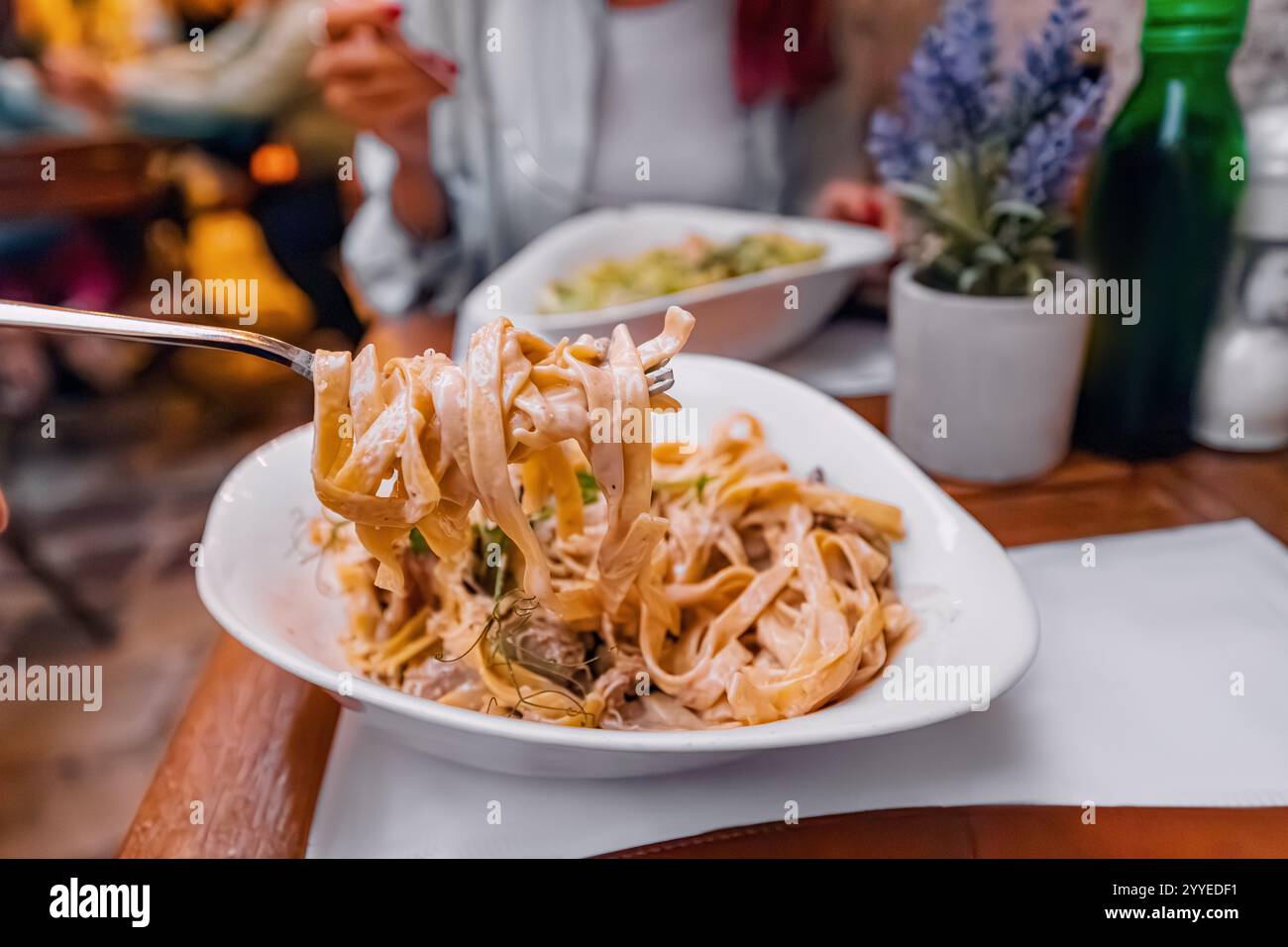 Tagliatelle cremose con salsa sollevata da una forchetta in un ristorante, con cene sfocate sullo sfondo Foto Stock