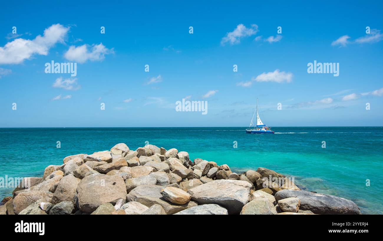 La barca a vela passa davanti a un affioramento roccioso, su acque turchesi, visto da una spiaggia privata a Noord lungo la costa di Aruba Foto Stock