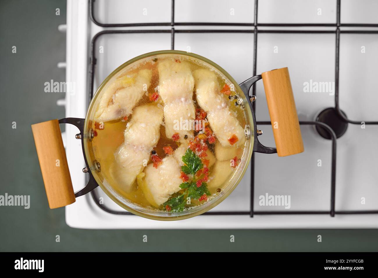 Una pentola di filetti di pesce bianco fiamma lentamente in brodo limpido, guarnita con erbe e spezie. La scena cattura un'atmosfera calda e invitante in cucina, em Foto Stock