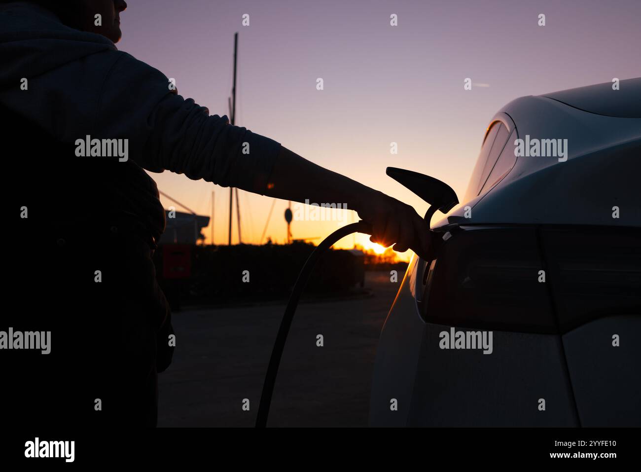 Una silhouette sorprendente di una persona che carica un'auto elettrica, con le vivaci sfumature di un tramonto sullo sfondo. Foto Stock