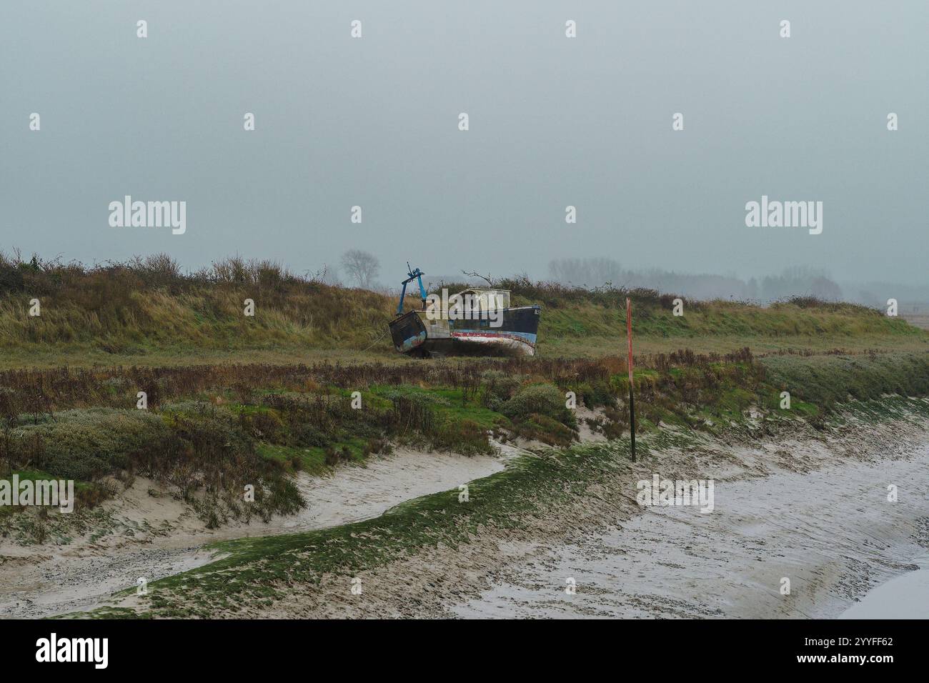 La barca da pesca abbandonata poggia su una riva fangosa sotto il cielo coperto vicino a praterie paludose Foto Stock