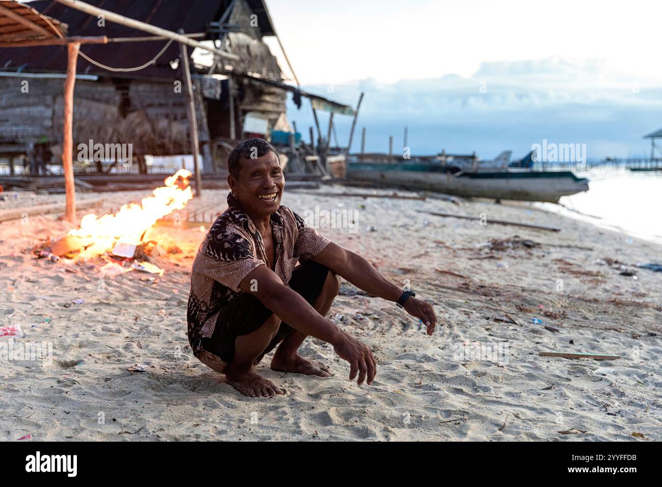 uomo della tribù bajau seduto su una spiaggia che brucia spazzatura di plastica che le correnti marine hanno portato sulla sua isola vicino all'isola di muna, sulawesi, indonesia Foto Stock