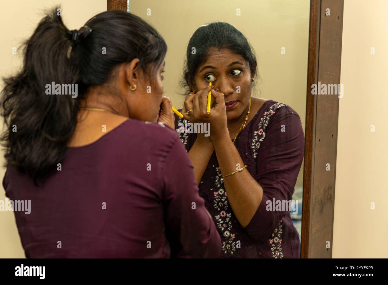 Una donna dell'Asia meridionale applica l'eyeliner, preparandosi per la sua giornata con un'espressione concentrata. Foto Stock