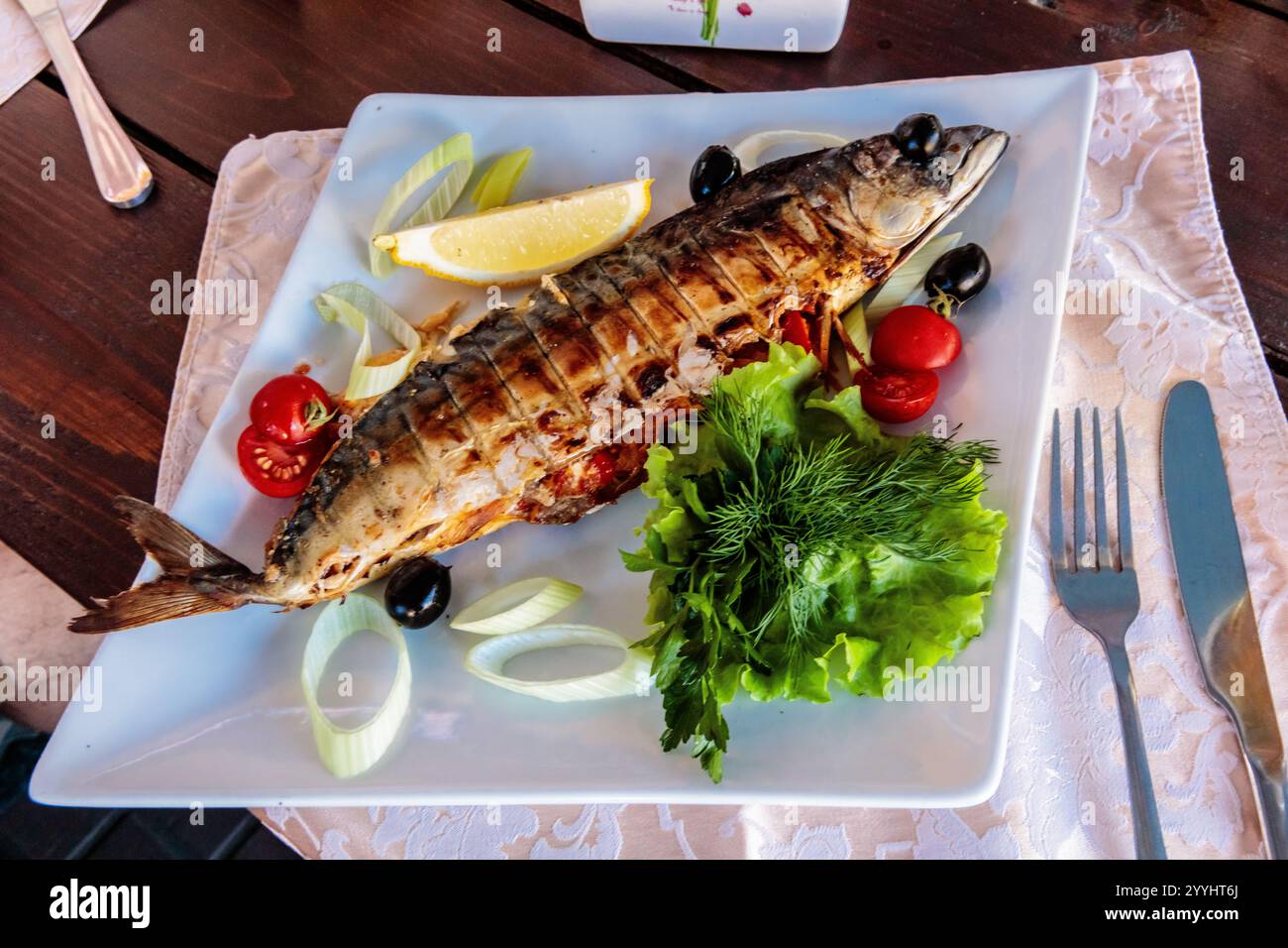 Un pesce viene servito su un piatto bianco con un contorno di verdure. Il pesce è grigliato e ha una fetta di limone sulla parte superiore. Le verdure includono lattuga, tomatoe Foto Stock