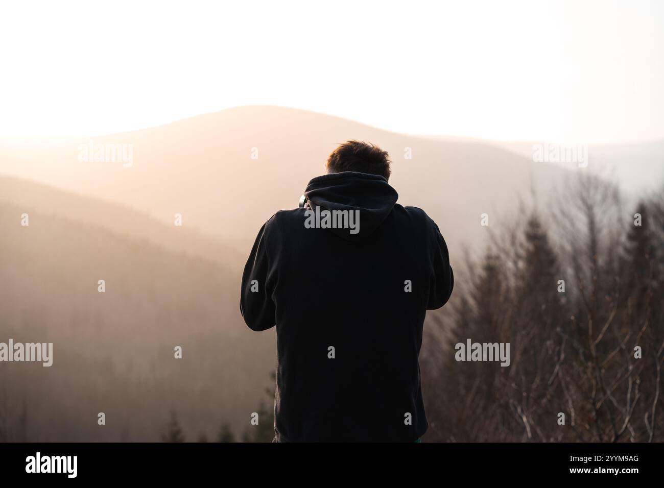 Un uomo che indossa una felpa con cappuccio nera si trova di fronte a una maestosa catena montuosa che viene catturata splendidamente alla luce del mattino Foto Stock