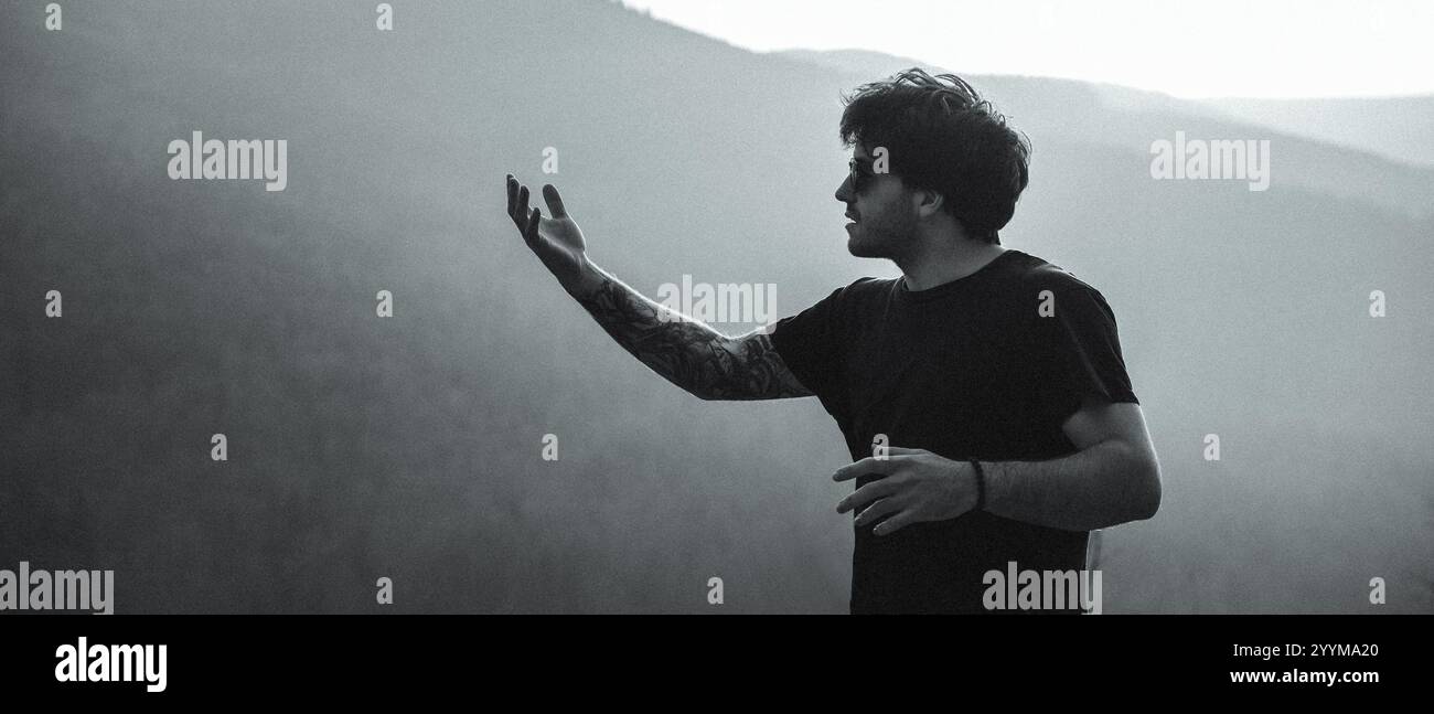 In un'accattivante foto in bianco e nero, un uomo vestito con una camicia nera si trova di fronte a un maestoso sfondo di montagna Foto Stock