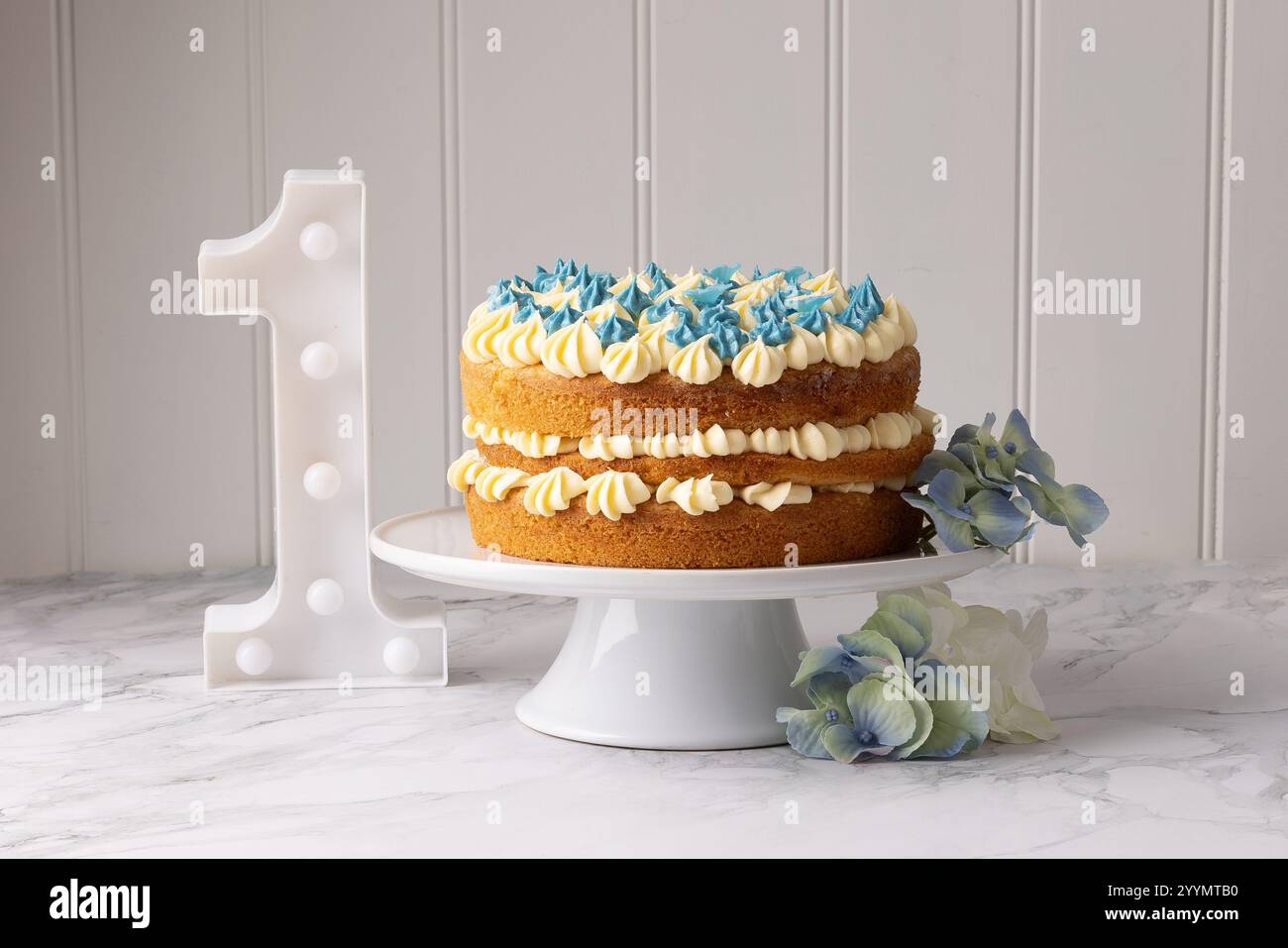 Una torta per festeggiare il primo compleanno, ghiacciata con crema di burro alla vaniglia e vortici blu, con un grande numero uno sul tavolo a lato. Foto Stock