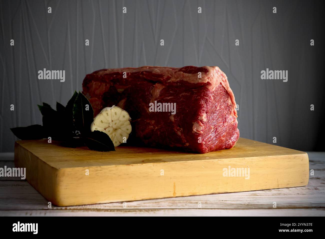 Un grosso taglio crudo crudo crudo o un giunto di filetto di manzo su una tavola rustica di legno con un bulbo d'aglio tagliato a croce e foglie di alloro. Foto Stock