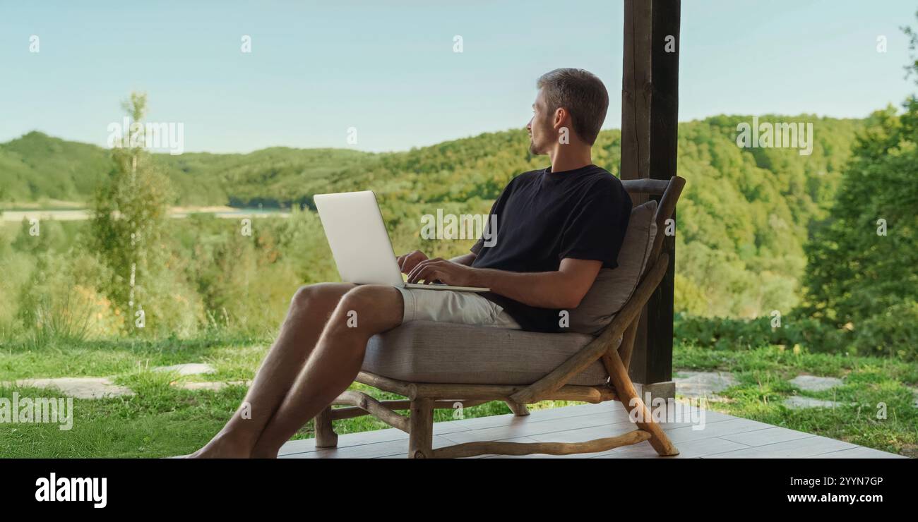 Giovane freelance maschio seduto su una sedia di legno, con laptop, che gode della natura e di una splendida vista sulla foresta mentre lavora in remoto. Equilibrio tra lavoro e vita privata. Copia spazio Foto Stock