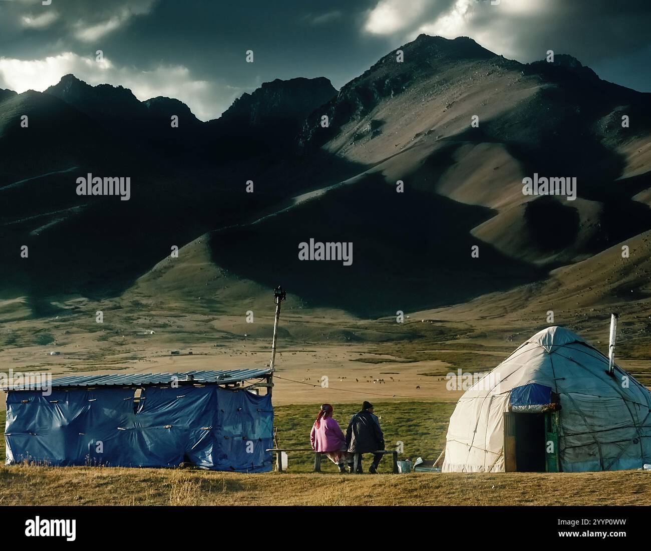 Due persone siedono davanti a una yurta e a una tenda contro un paesaggio di montagna, il cielo del tramonto proietta ombre sulla scena. Foto Stock