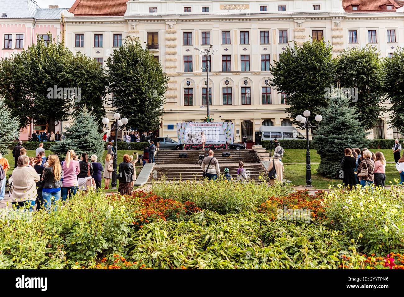 Una grande folla di persone è riunita di fronte a un edificio con un palco. La gente è in piedi in un'area giardino con fiori e alberi. La scena è li Foto Stock