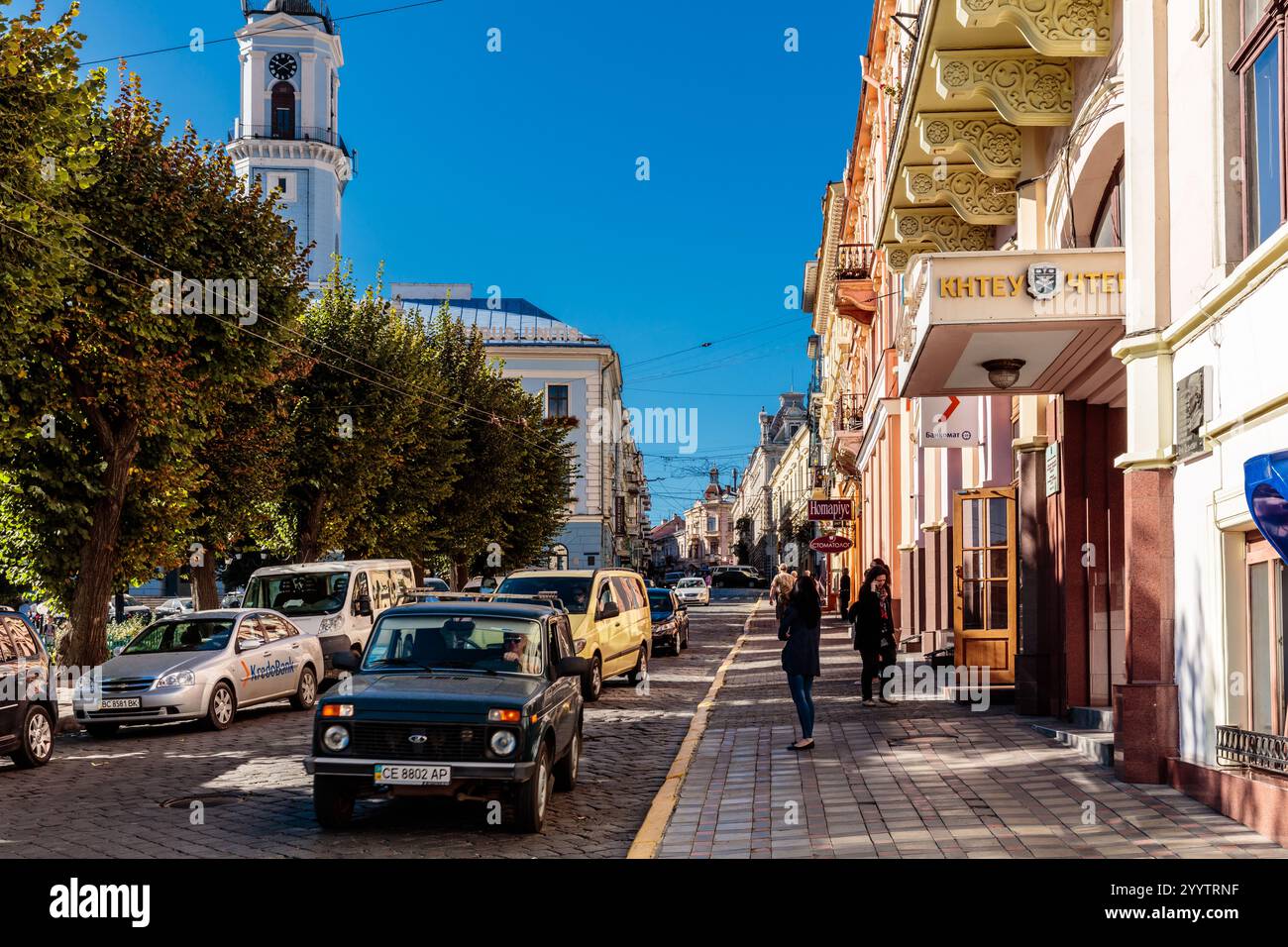 Una strada trafficata con un grande edificio sullo sfondo. La gente cammina lungo la strada e le auto sono parcheggiate lungo il lato Foto Stock