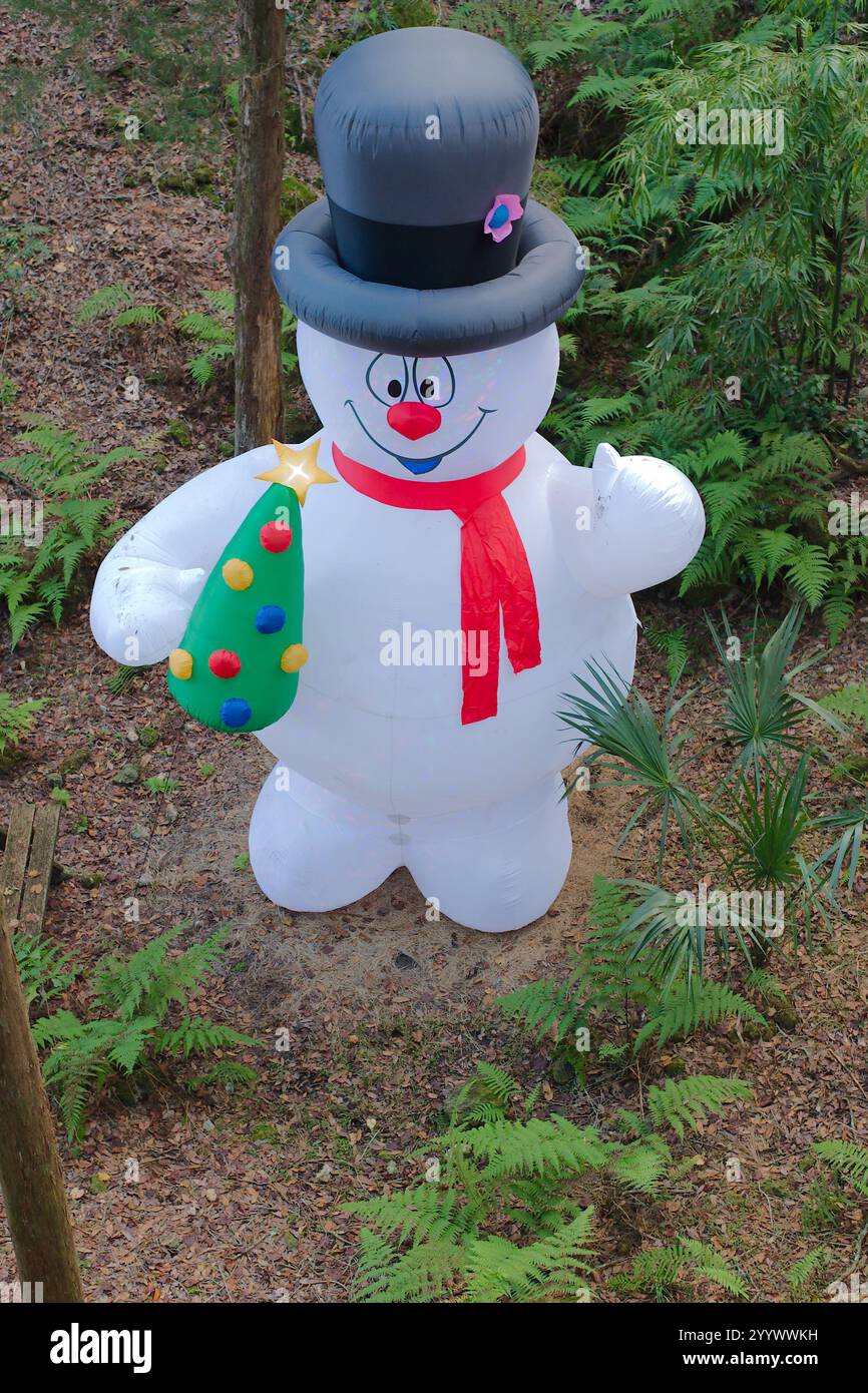 Pupazzo di neve gonfiabile vestito per Natale. Indossa un cappello, vestiti rossi e neri. Erba verde, foglie marroni e alberi sullo sfondo. Foto Stock