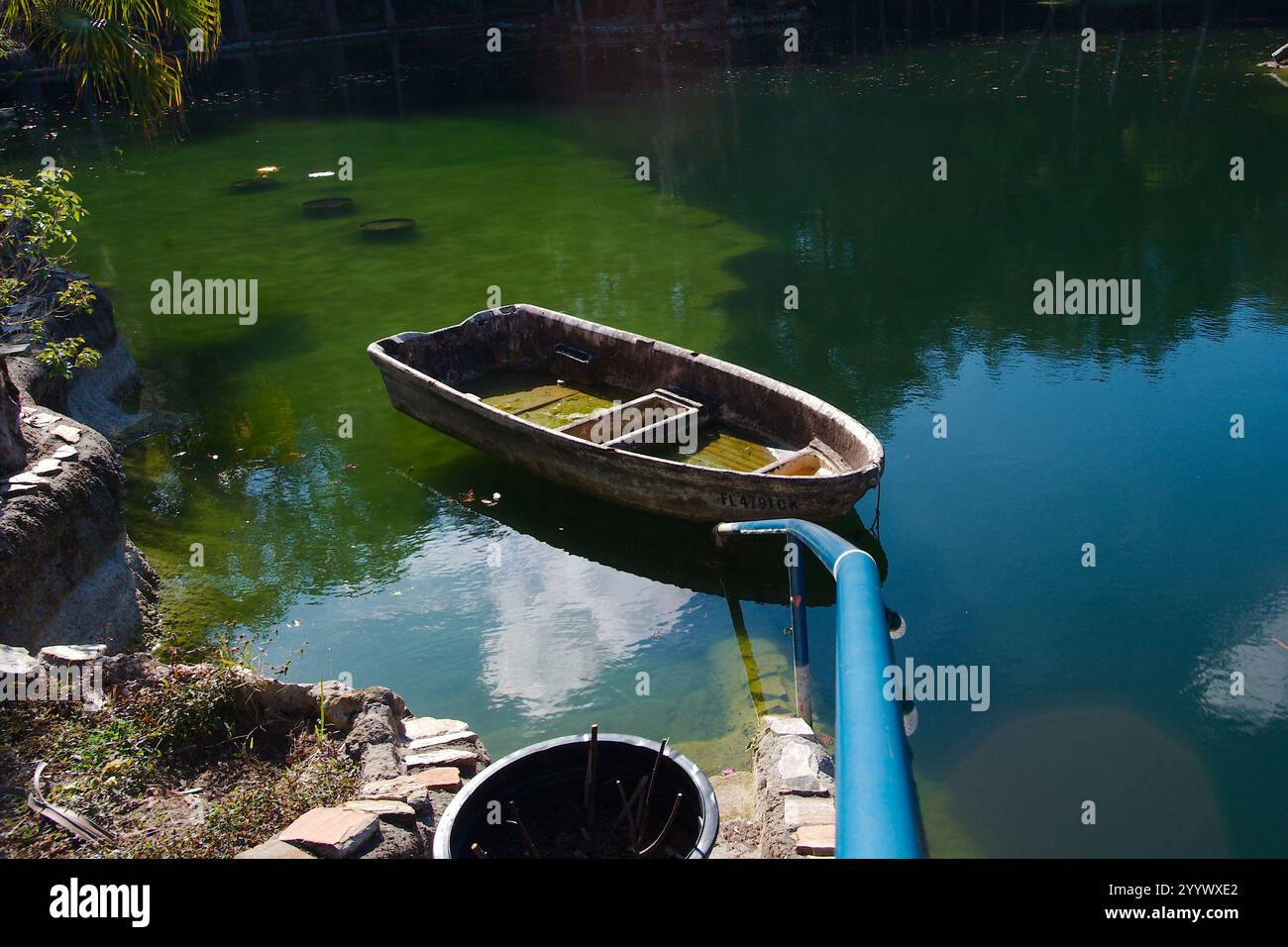 Le linee che scendono lungo un corrimano blu fino a una vecchia barca a remi con pietre sul lato. Riflessi del cielo e delle nuvole in acque blu e verdi. Su un Foto Stock