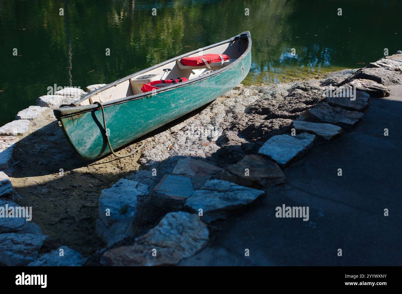 Vecchia canoa verde graffiata incorniciata tra pietre sul bordo dell'acqua, corda appesa e giubbotti di salvataggio all'interno. Riflessi del cielo e delle nuvole in calmo smo Foto Stock