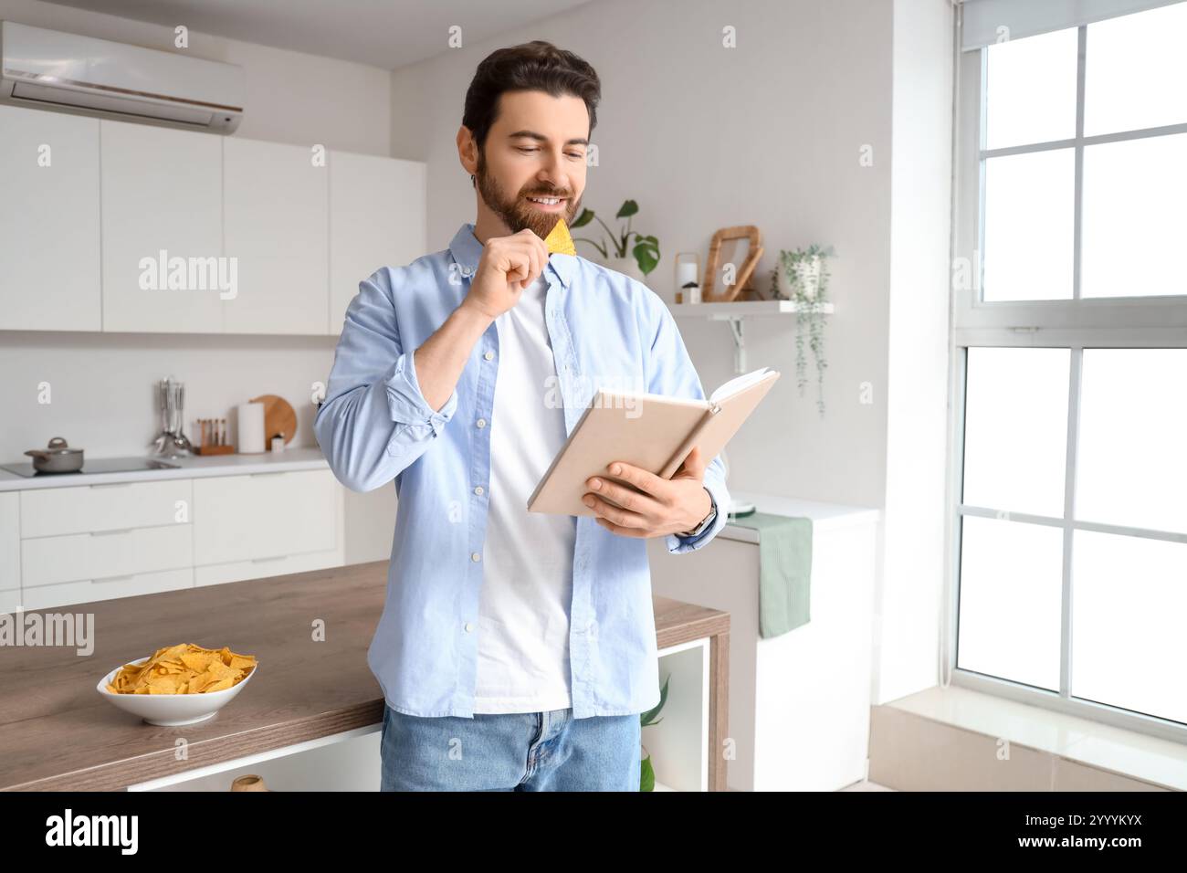 Uomo che mangia gustosi nachos e legge un libro in cucina Foto Stock