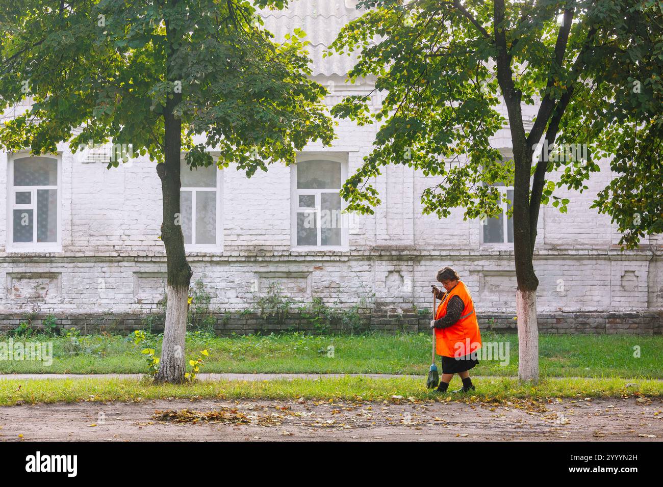 Lavoratore municipale in gilet arancione che spazza foglie cadute vicino a edifici in mattoni bianchi e alberi. Scenario di manutenzione urbana autunnale. Progettazione per editoriale e c Foto Stock