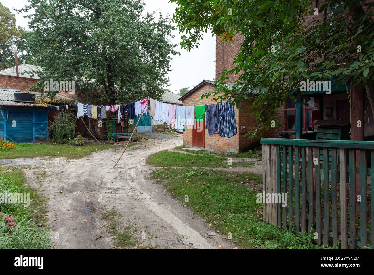 Vestiti che si asciugano su una stoffa in un cortile rurale con case in mattoni e vegetazione. Una scena all'aperto della vita di tutti i giorni. Progettazione destinata a essere utilizzata in materiali editoriali, Foto Stock