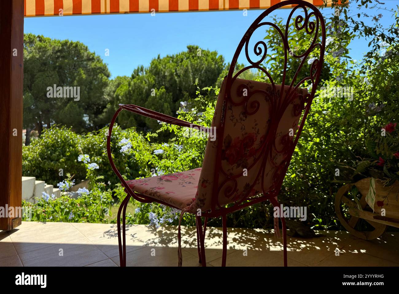 Classica sedia rossa in ferro battuto e cuscini rosa rosso vecchio stile sulla terrazza della villa vacanze nel giardino, alberi, fiori, cielo blu Foto Stock