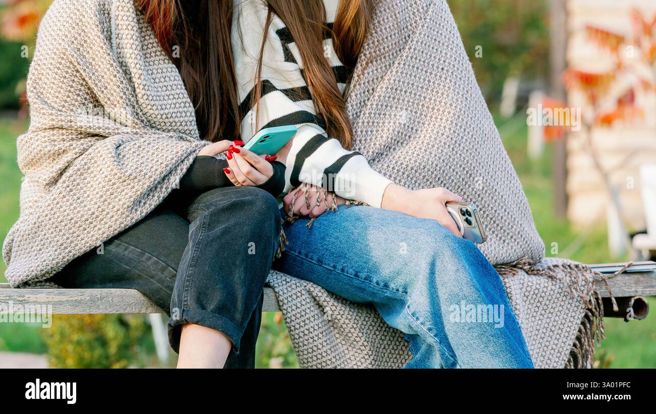 Due giovani donne sedute insieme all'aperto, avvolte in una coperta accogliente, che tengono in mano gli smartphone e si godono un momento informale con il bel tempo. Foto Stock Due giovani donne sedute insieme all'aperto, avvolte in una coperta accogliente, che tengono in mano gli smartphone e si godono un momento informale con il bel tempo. Foto Stock