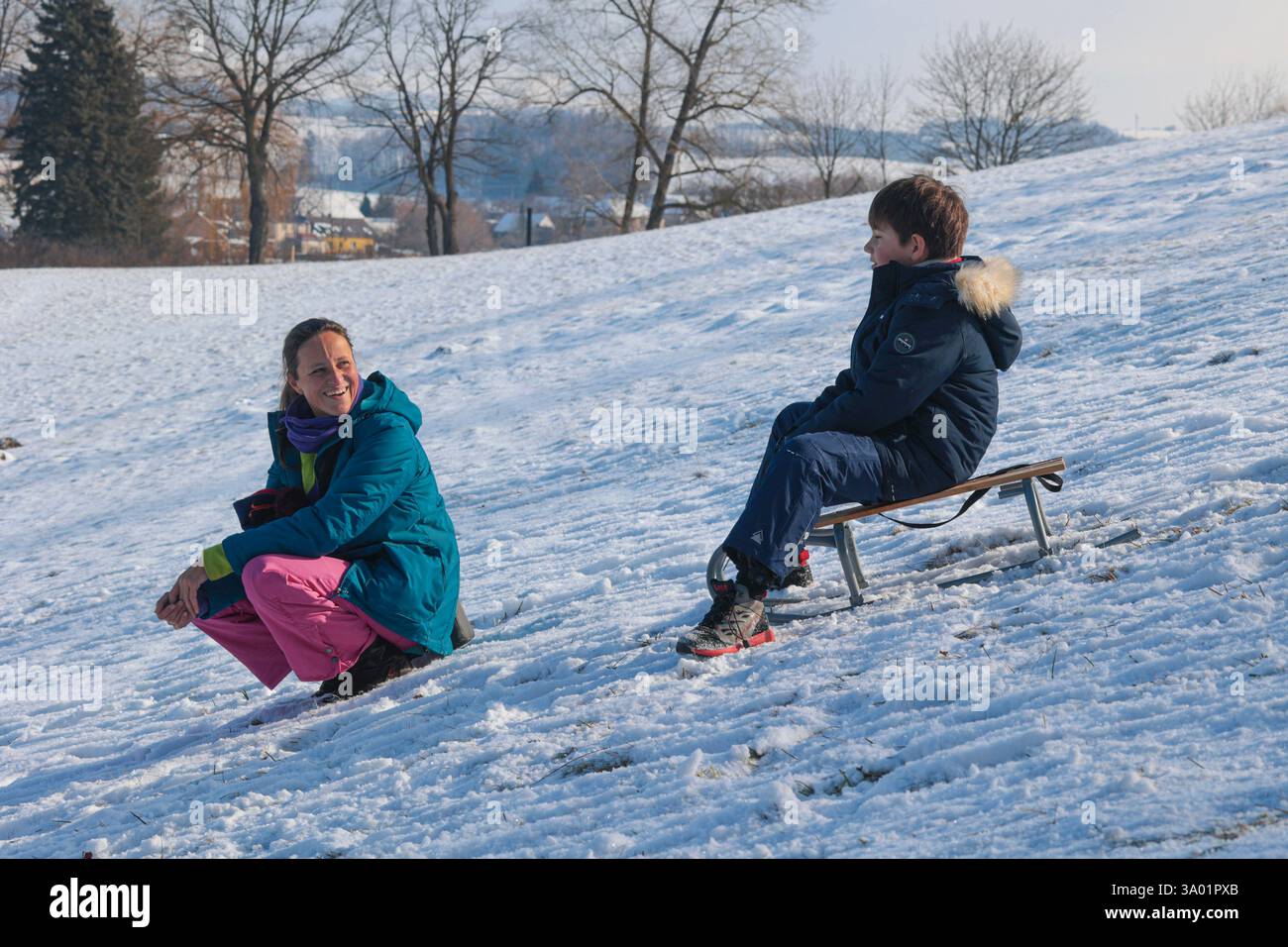 Donna ridendo che guarda un ragazzo che slitta su una collina innevata Foto Stock