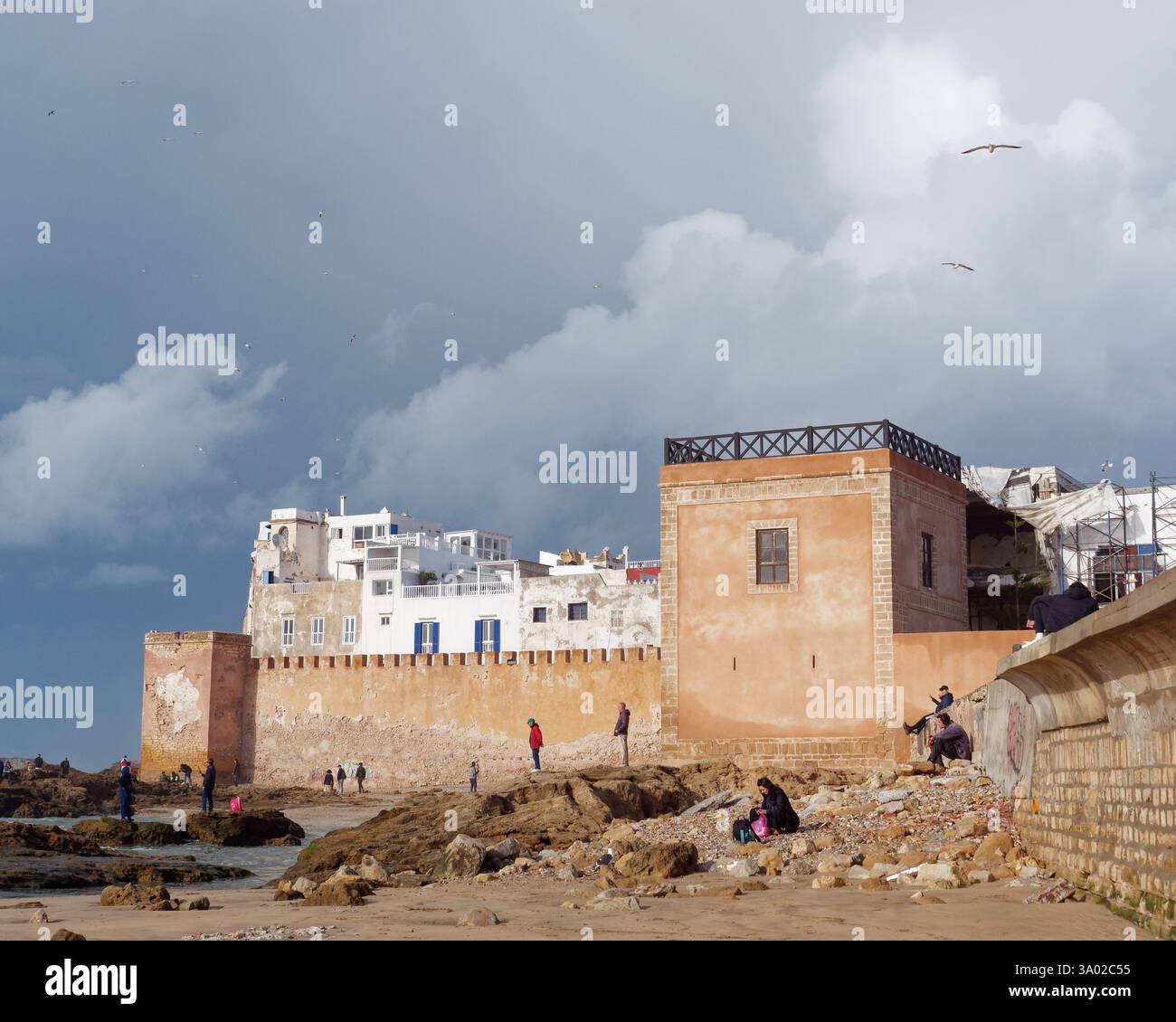 Persone su una spiaggia rocciosa con medina storica alle spalle e cieli drammatici a Essaouria, Marocco. Febbraio/marzo 2025 Foto Stock