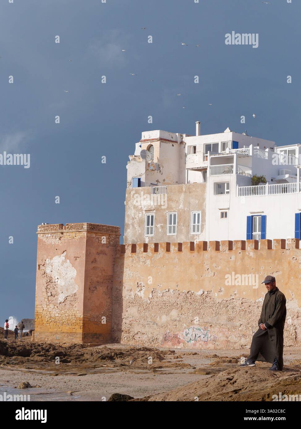 Un uomo del posto si erge su una spiaggia rocciosa con medina storica alle spalle e cieli drammatici a Essaouria, Marocco. Febbraio/marzo 2025 Foto Stock