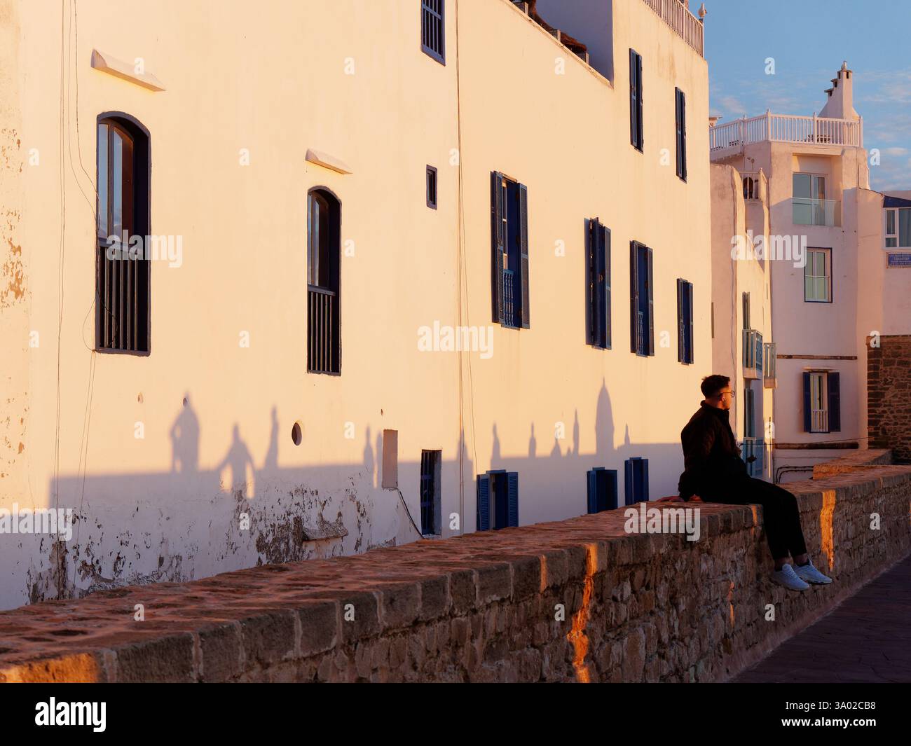 L'uomo siede sulle mura interne della città della Medina mentre le ombre sono gettate sul muro dietro la luce della sera a Essaouria, in Marocco. Febbraio/marzo 2025 Foto Stock