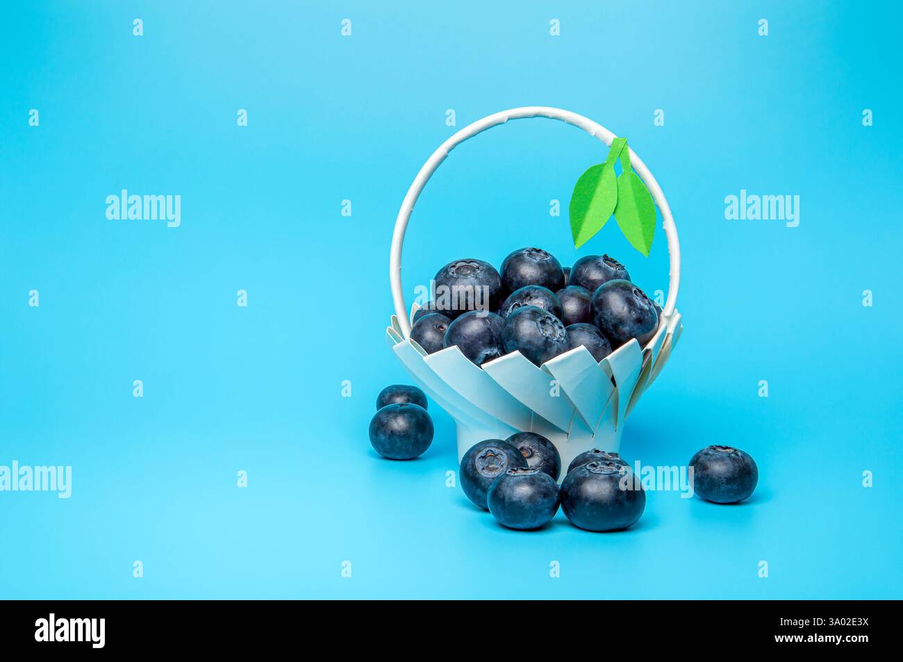 Mirtilli freschi crudi in un cestino di carta bianca fatto in casa con foglie verdi su sfondo blu. Foto Stock