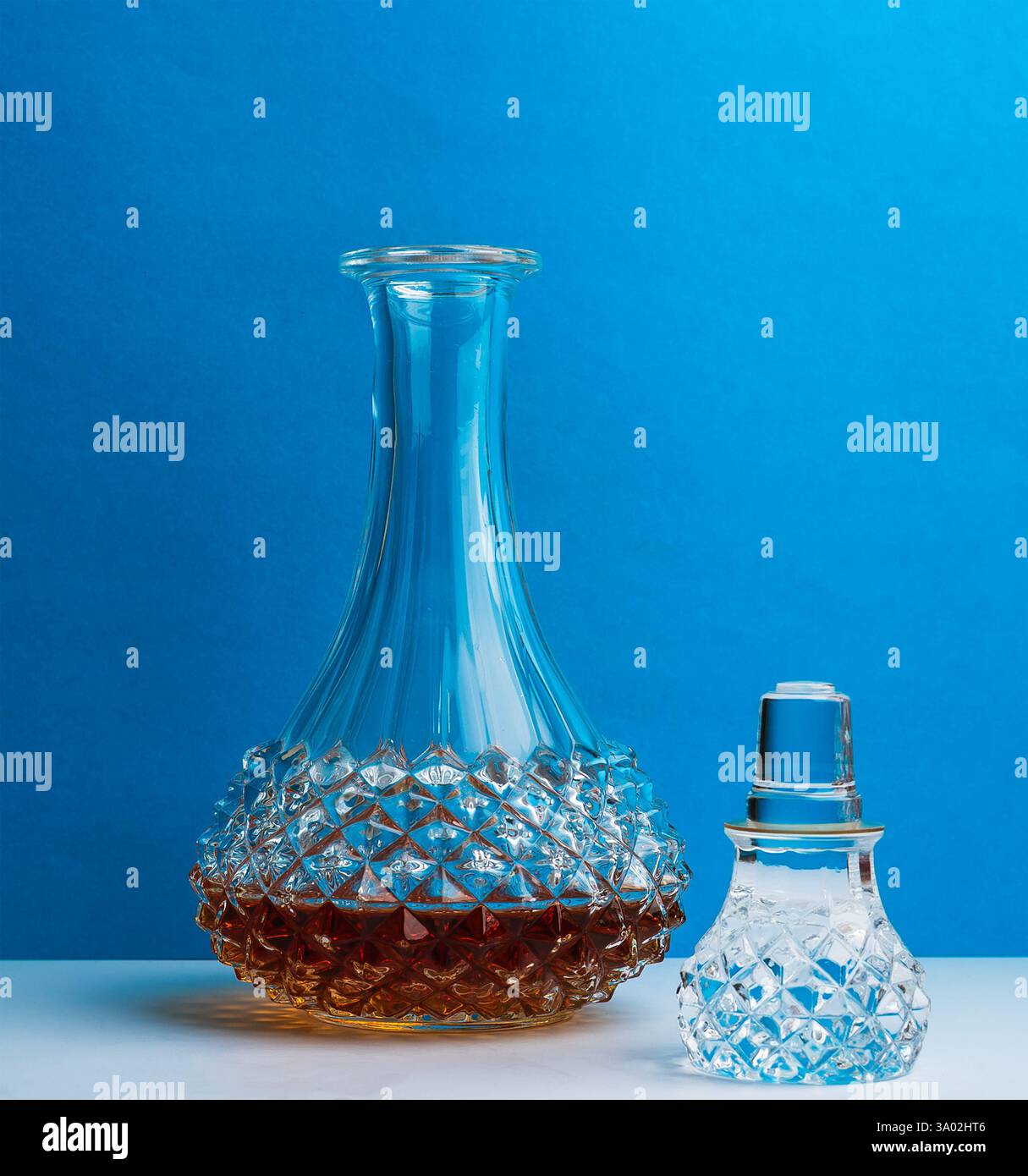 Un decanter in vetro finemente lavorato e un tappo coordinato sono posizionati su una superficie, rivelando un liquido color ambra profondo sotto uno sfondo blu. Foto Stock