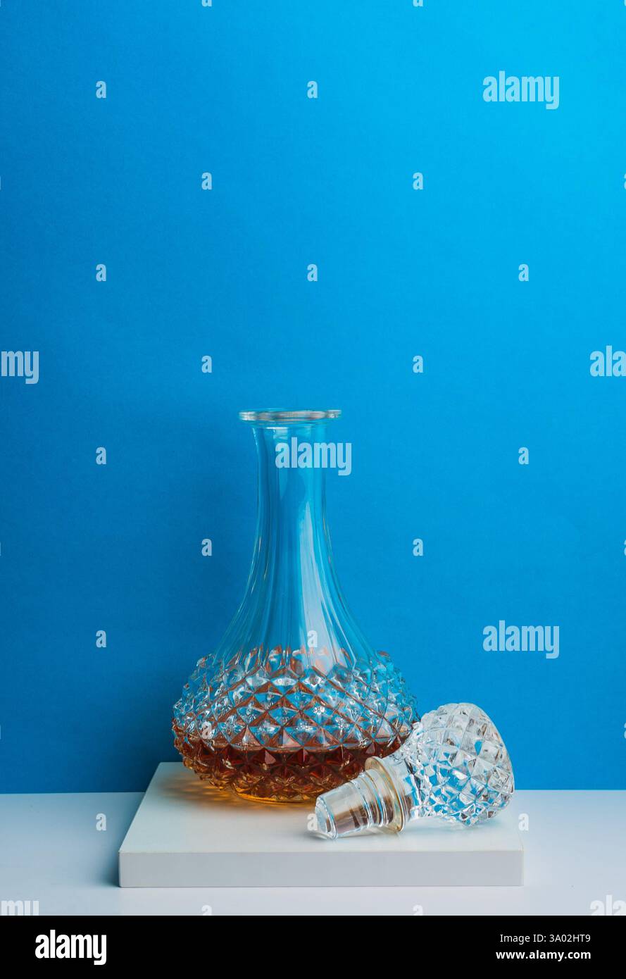 Un decanter in cristallo di ottima fattura e un tappo coordinato poggiano su una semplice piattaforma bianca, su uno sfondo blu vibrante che mette in risalto il loro aspetto Foto Stock