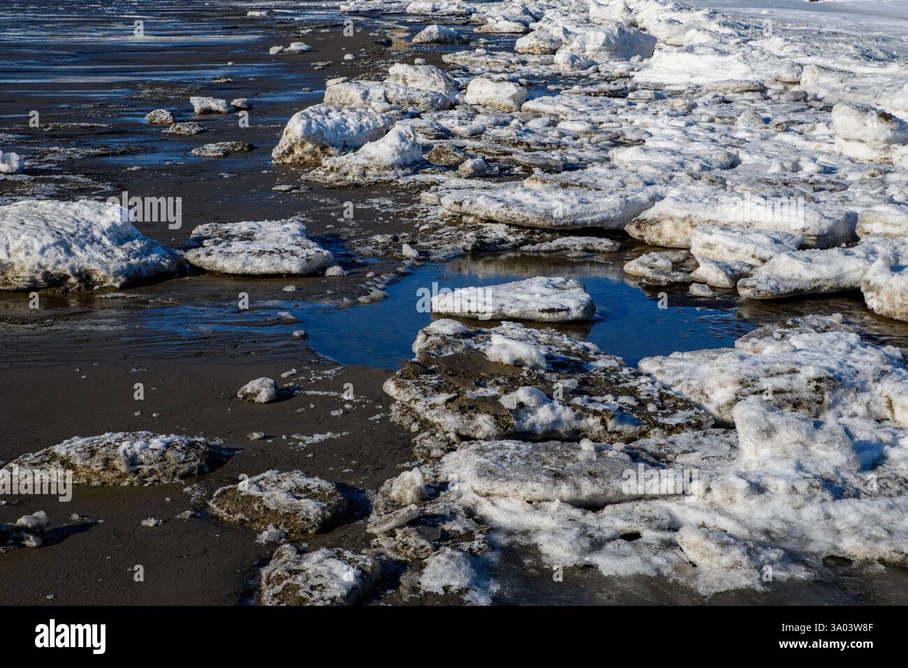 Blocchi di ghiaccio rotto riposano su una spiaggia bagnata e sabbiosa. Il ghiaccio è sporco. Il cielo blu si riflette nelle pozzanghere. Foto Stock