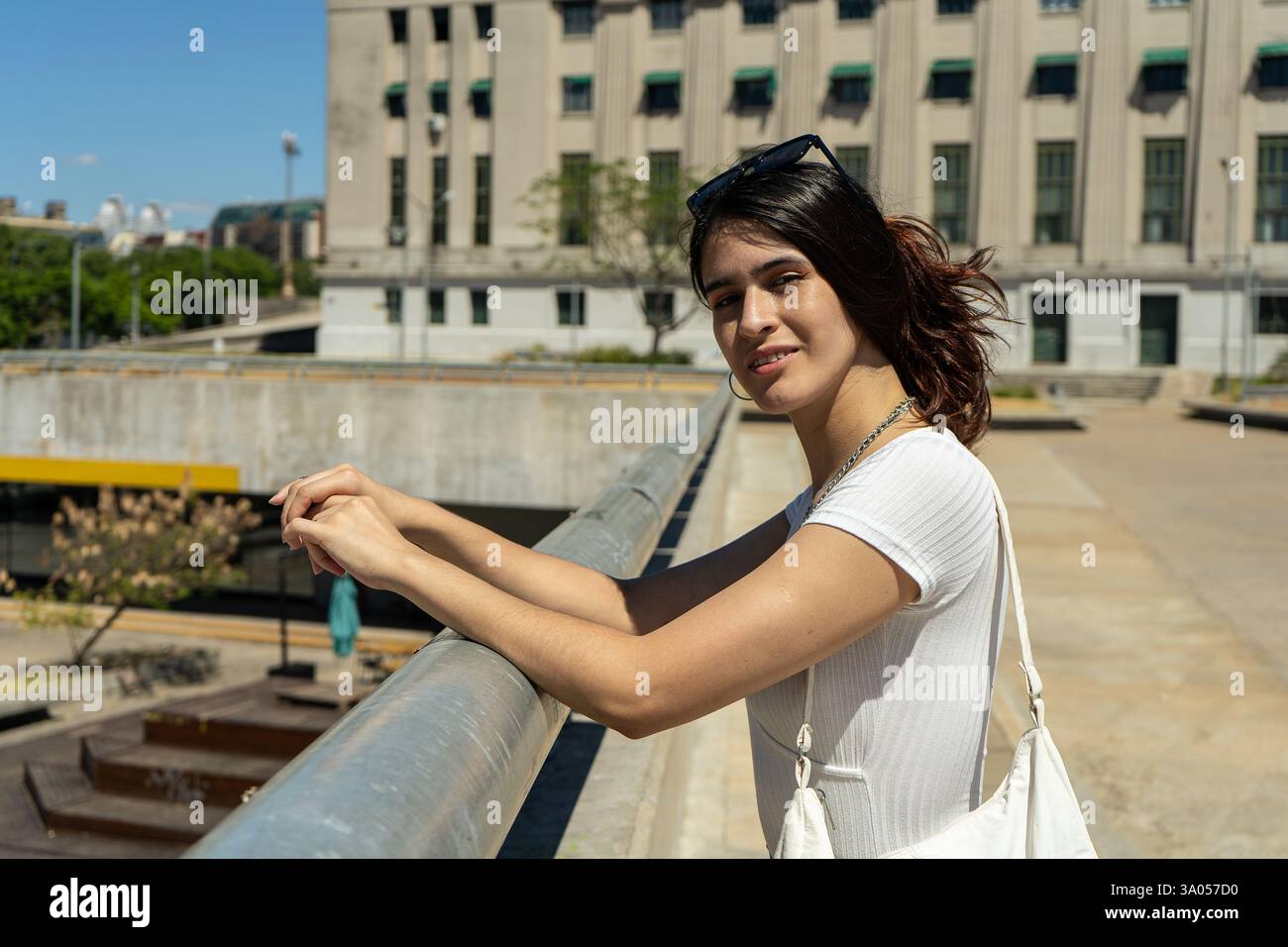 Giovane donna latina elegante che gode di una giornata di sole in città, appoggiata su una ringhiera con una posa sicura e rilassata Foto Stock