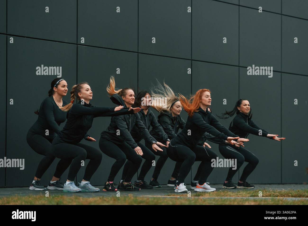 Fare degli squat. Un gruppo di donne sportive si trova all'aperto vicino all'edificio nero. Foto Stock