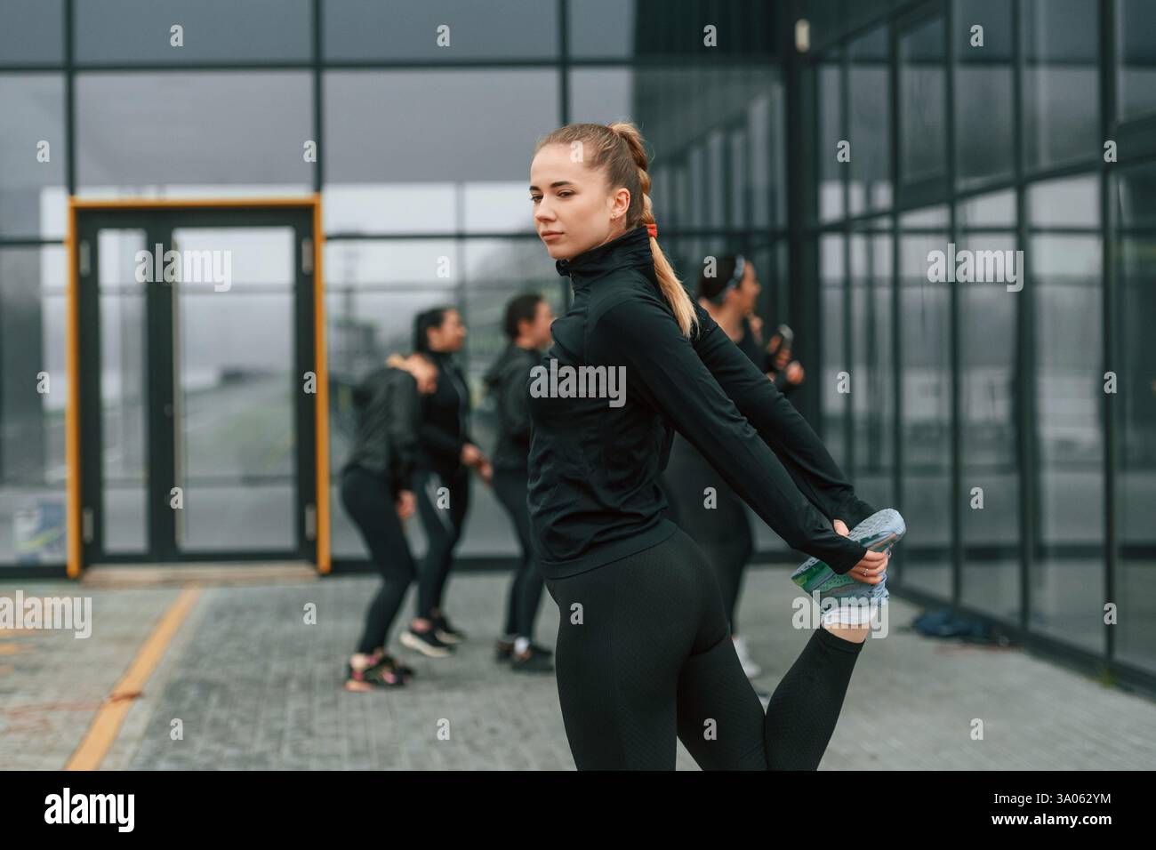 La bella donna è in piedi davanti ai suoi amici. Un gruppo di donne sportive si trova all'aperto vicino all'edificio nero. Foto Stock