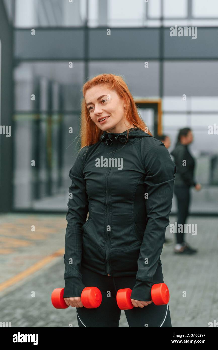 La bella donna è in piedi davanti ai suoi amici. Un gruppo di donne sportive si trova all'aperto vicino all'edificio nero. Foto Stock
