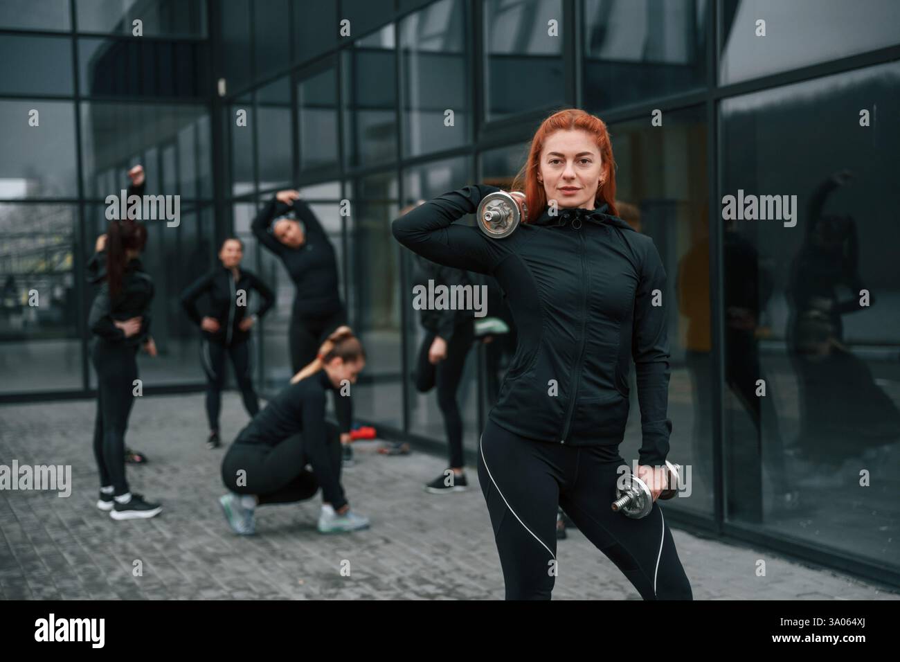 Sollevamento manubri. Un gruppo di donne sportive si trova all'aperto vicino all'edificio nero. Foto Stock