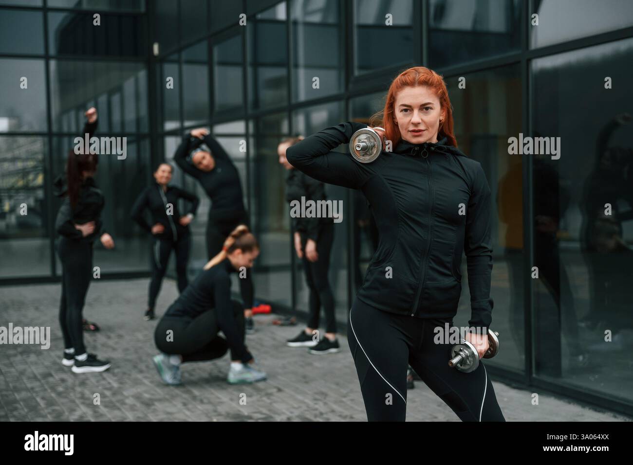 Sollevamento manubri. Un gruppo di donne sportive si trova all'aperto vicino all'edificio nero. Foto Stock