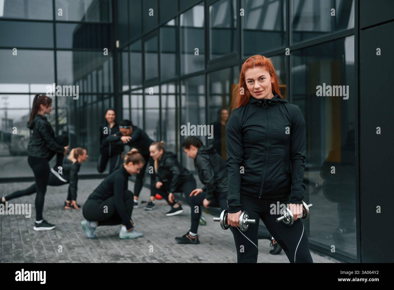 Sollevamento manubri. Un gruppo di donne sportive si trova all'aperto vicino all'edificio nero. Foto Stock