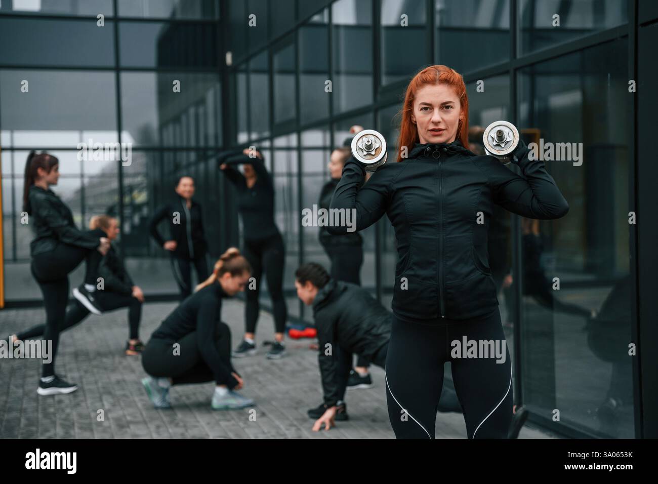 La bella donna è in piedi davanti ai suoi amici. Un gruppo di donne sportive si trova all'aperto vicino all'edificio nero. Foto Stock